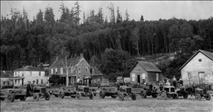 A row of tractors in front of a row of houses with forest behind them. There are men on the tractors. 